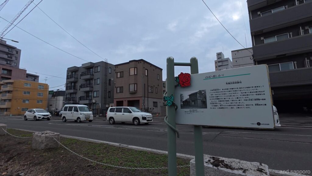 初春の歩道脇に立つ「有島武郎邸跡」の解説板。有島の肖像写真と、かつてここに建っていた寄宿舎（有島の住居）の説明が書かれている。